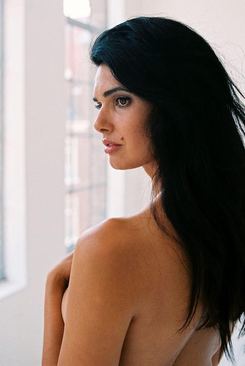 A woman with long dark hair looks over her shoulder towards a window.