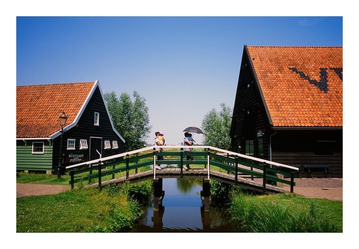 Two people stand on a green bridge over a canal between two traditional Dutch houses with orange roofs.