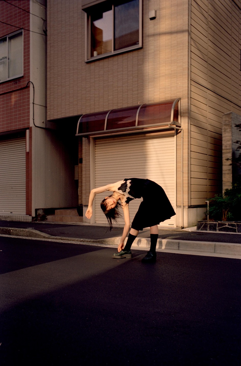 A woman in a black dress bends over on a street next to a building with garage doors.