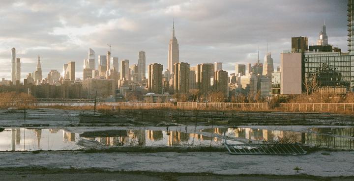 A cityscape with tall buildings is reflected in puddles on a wasteland under a cloudy sky.