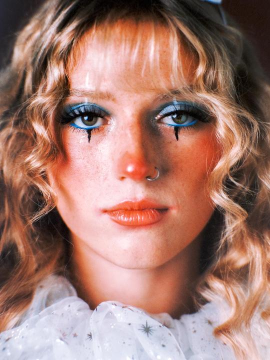 A woman with curly blonde hair and clown makeup looks directly at the viewer.
