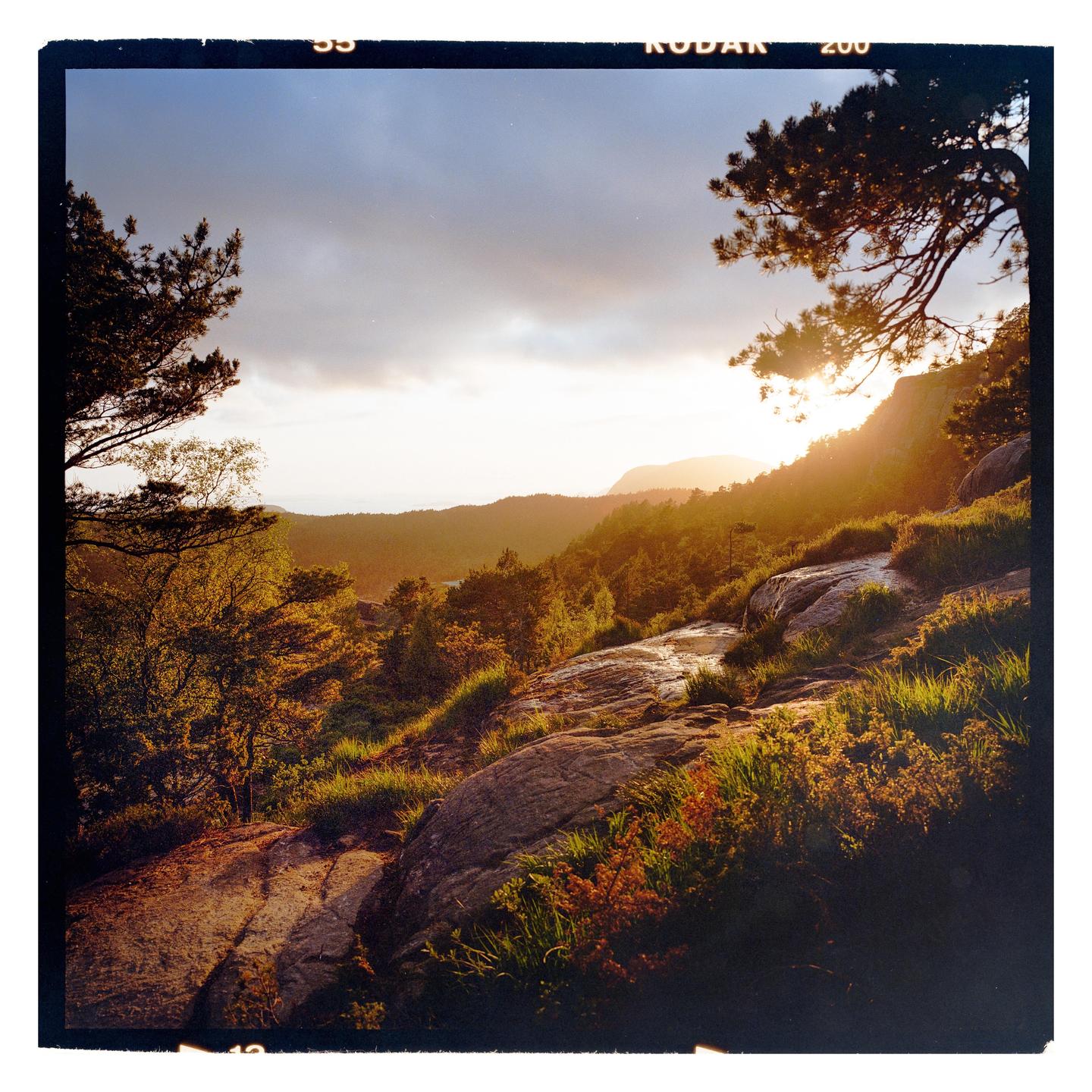 The sun sets over a mountainous landscape with trees and rocky terrain.