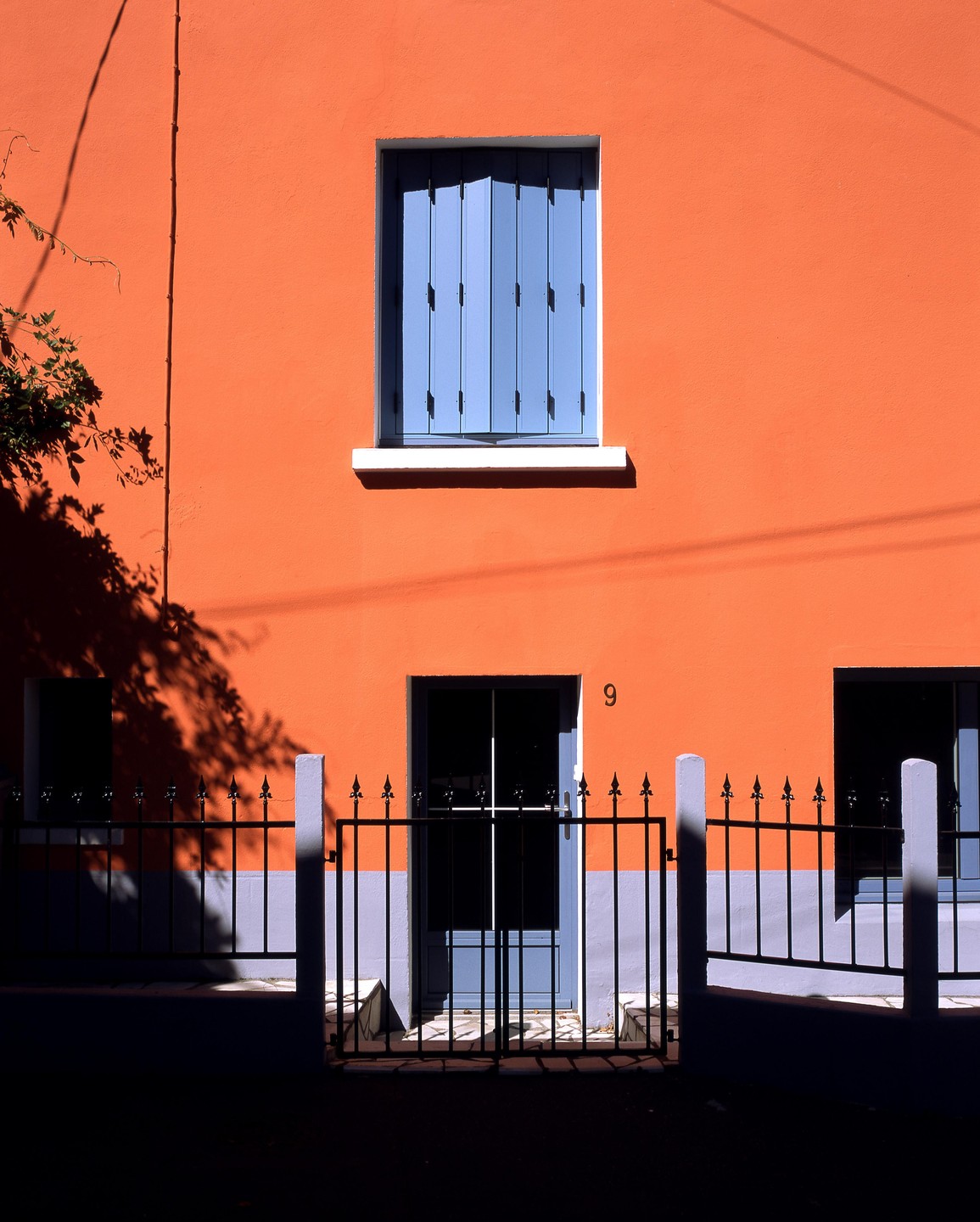 A bright orange building with blue shutters and a black fence is illuminated by strong sunlight.