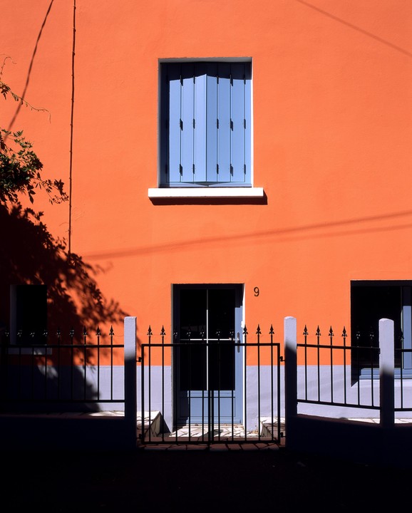 A bright orange building with blue shutters and a black fence is illuminated by strong sunlight.