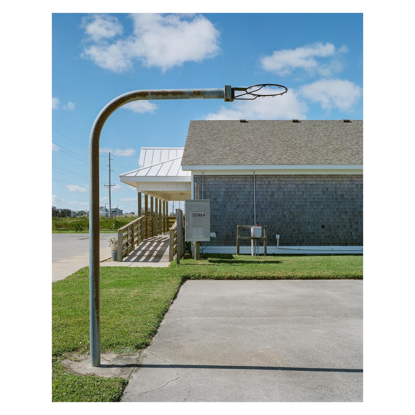 A rusted basketball hoop stands in front of a building with a shingled wall and a white roof under a blue sky with clouds.