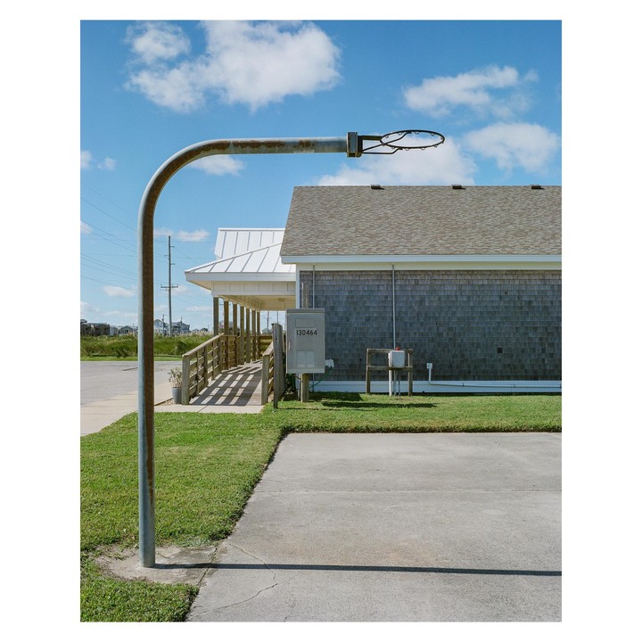 A rusted basketball hoop stands in front of a building with a shingled wall and a white roof under a blue sky with clouds.