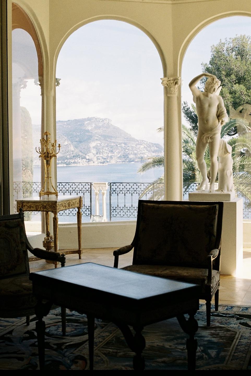 A room with arched windows overlooks a bay with mountains in the distance, with a statue of a man and a dog on the right.