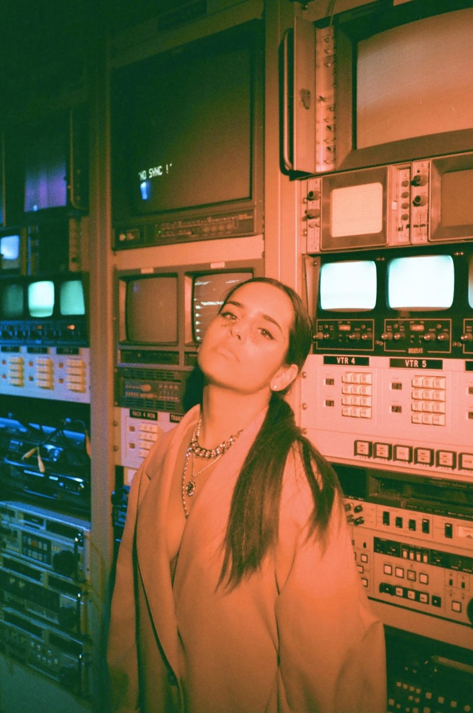 A woman with dark hair and a blazer stands in front of a wall of old electronic equipment and monitors.