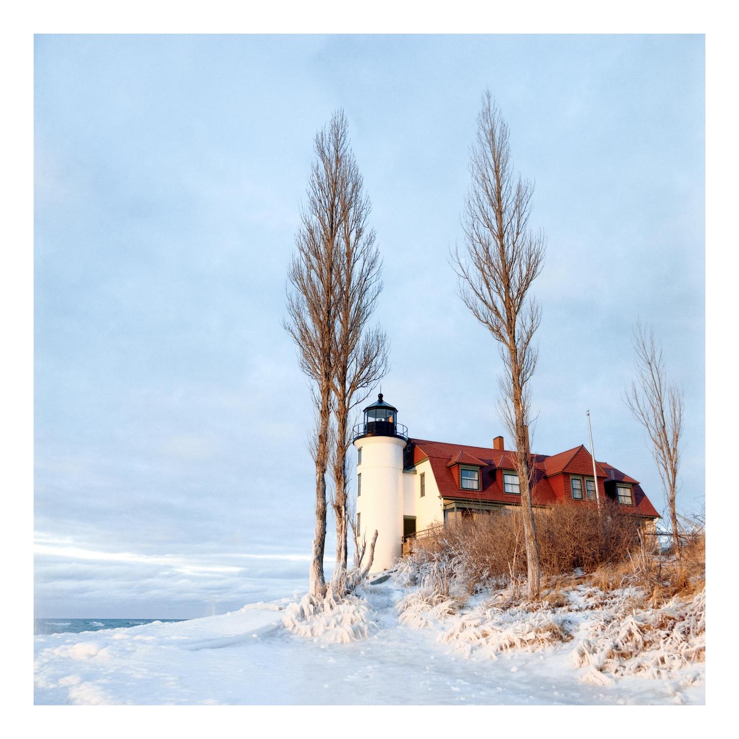 A white lighthouse with a red roof stands on a snowy hill next to a body of water under a pale sky.