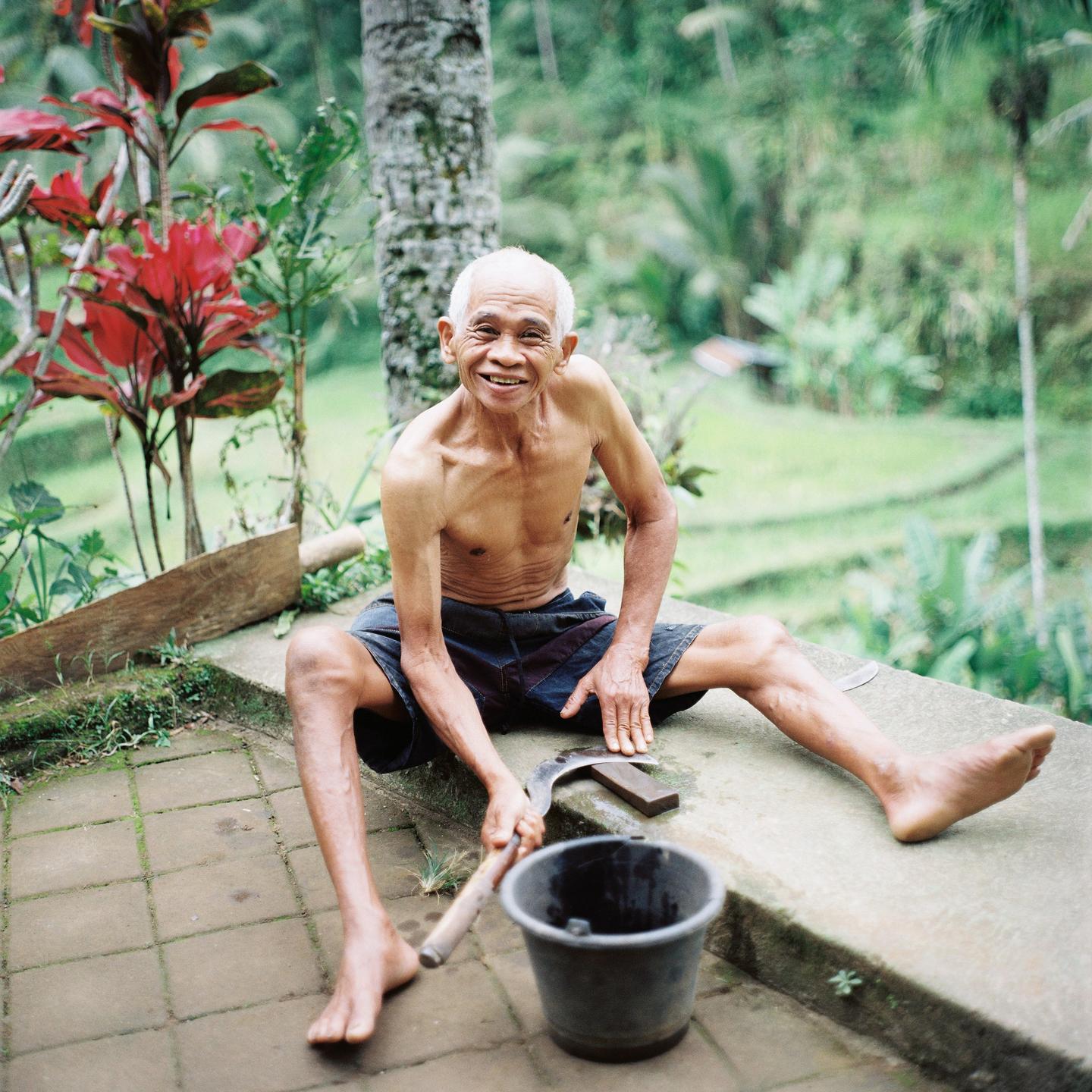 A man sits shirtless on a concrete patio, holding a sickle and smiling at the viewer.