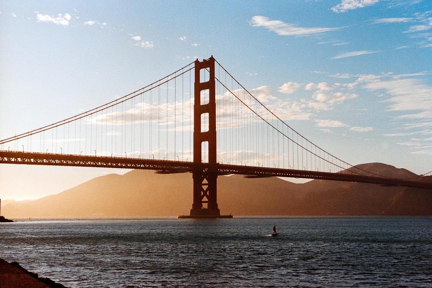 A person on a surfboard is in the water in front of a large suspension bridge with mountains in the background.