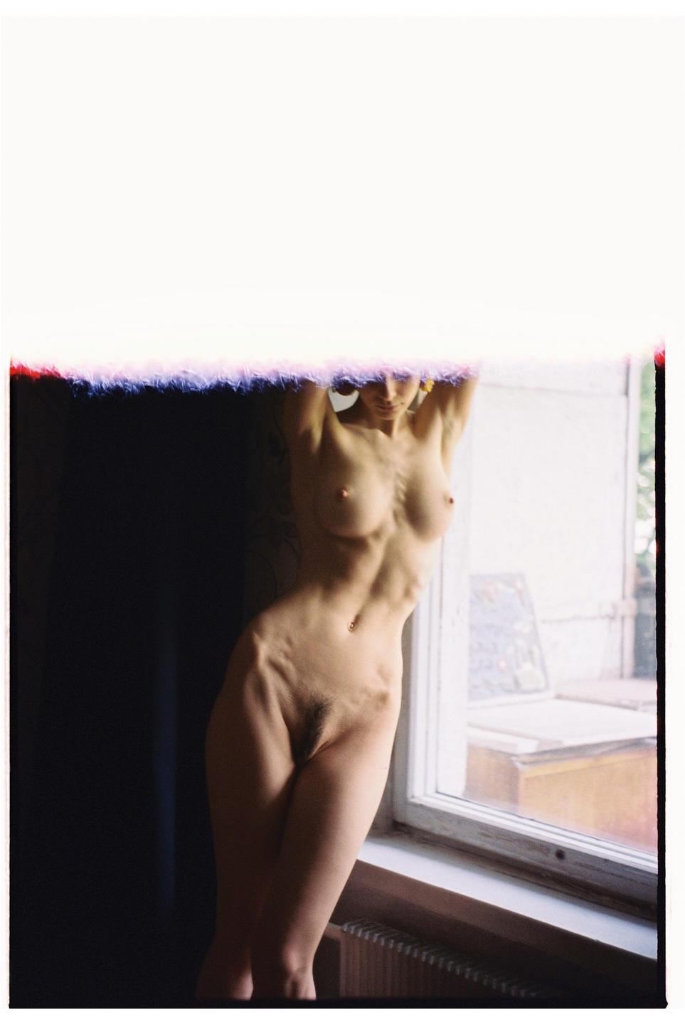 A nude woman stands with her arms raised, leaning against a wall next to a window.