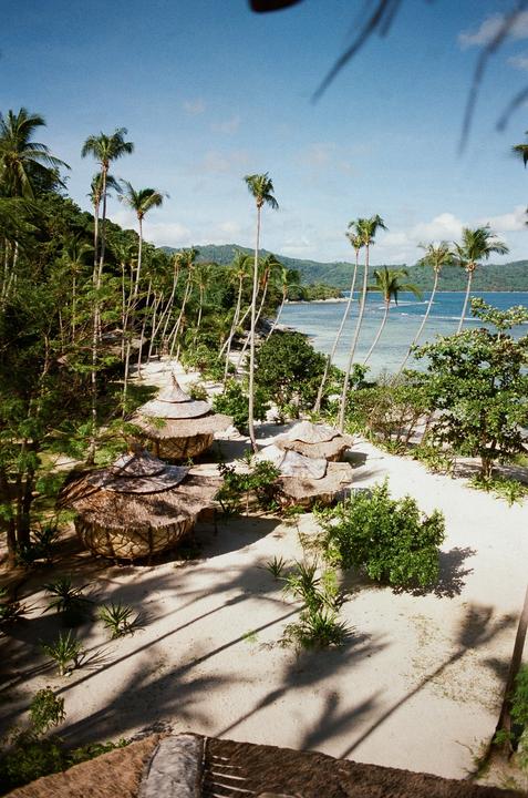 A tropical beach with palm trees, thatched-roof huts, and mountains in the background is shown from an elevated perspective.
