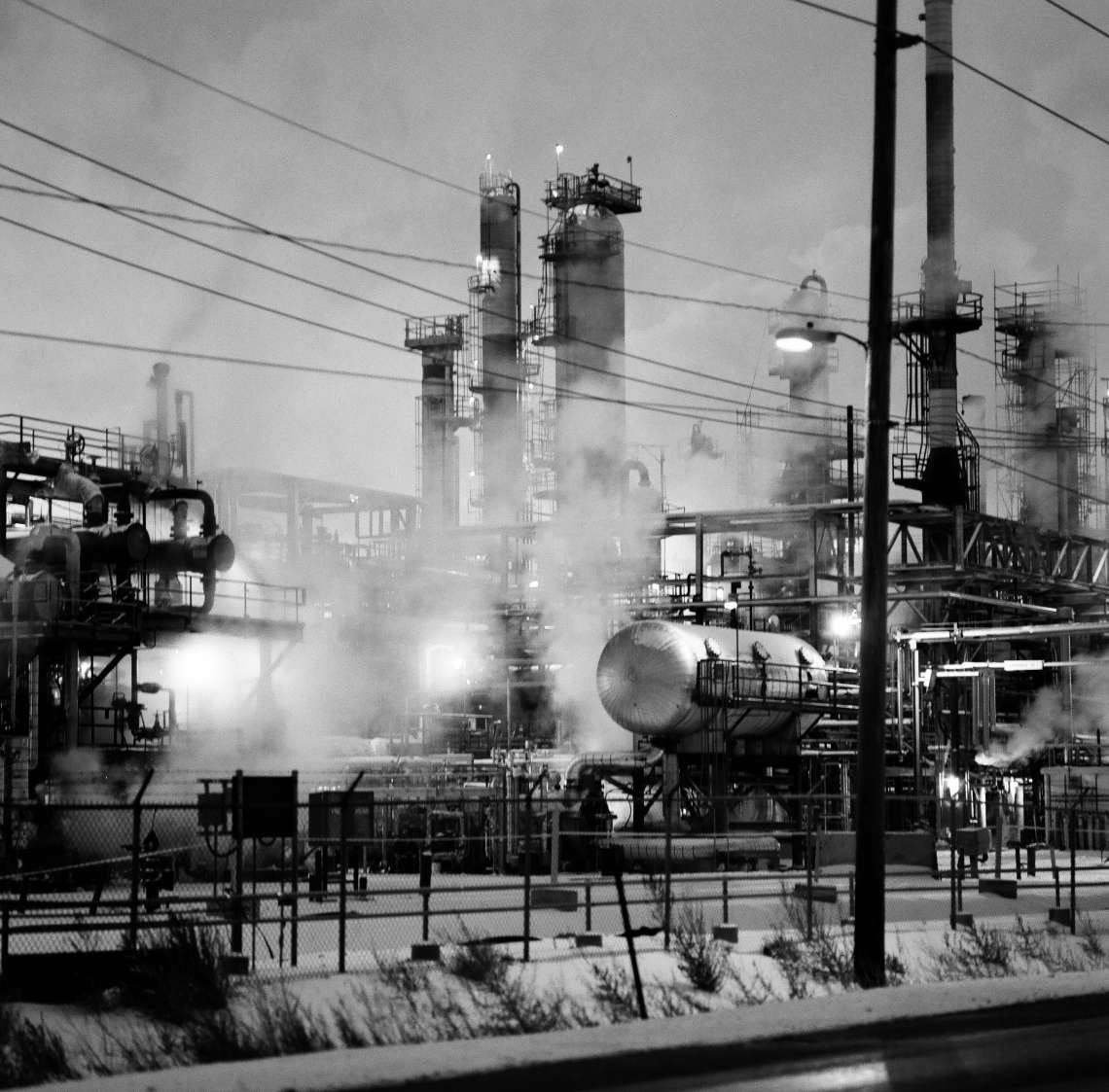 A large industrial complex with multiple towers, pipes, and tanks emits steam into the night sky, with power lines and a fence in the foreground.