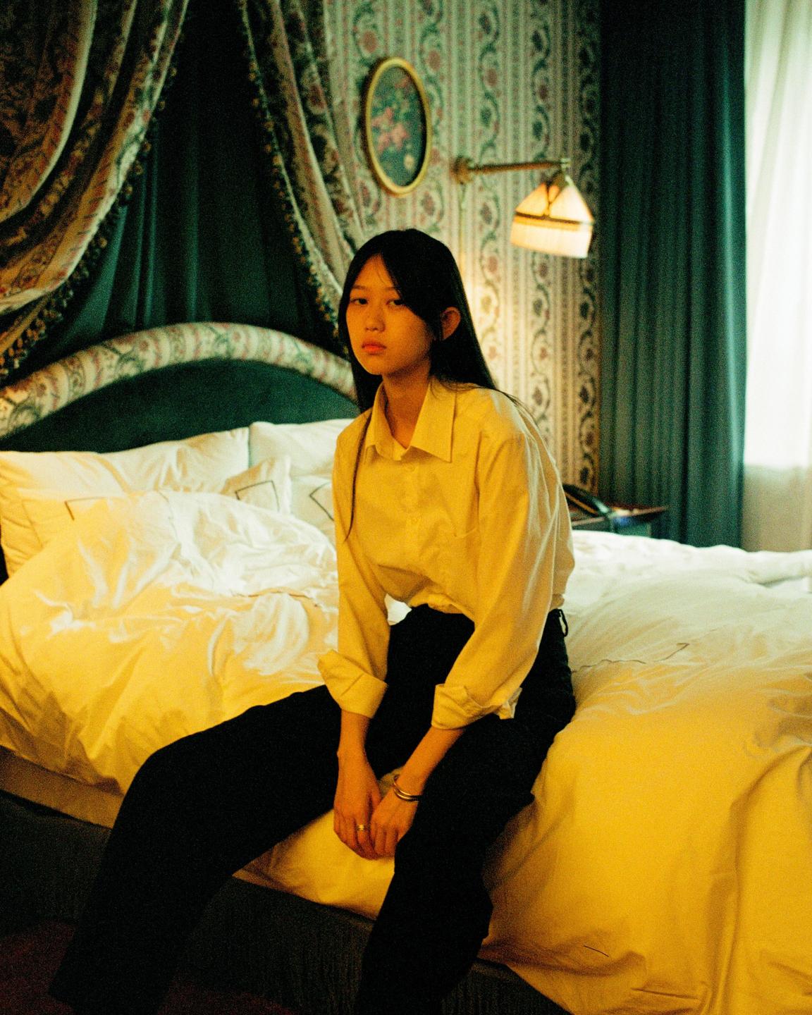 A woman sits on a bed in a hotel room with patterned wallpaper and a canopy bed.