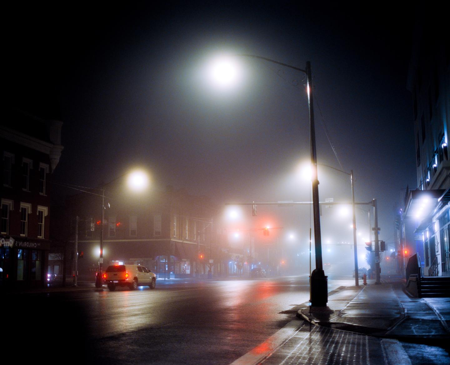 A foggy street at night is illuminated by streetlights and traffic lights, with a vehicle driving away from the viewer.
