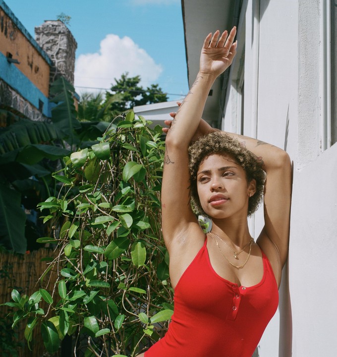 A woman in a red top leans against a white wall with green foliage and a building in the background.