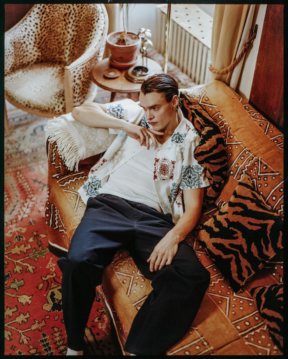 A man wearing a patterned shirt and dark pants reclines on a patterned sofa next to a leopard print chair.