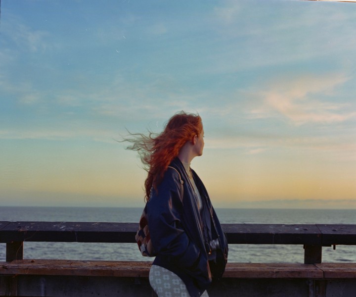 A woman with red hair stands on a pier looking out at the ocean at sunset.