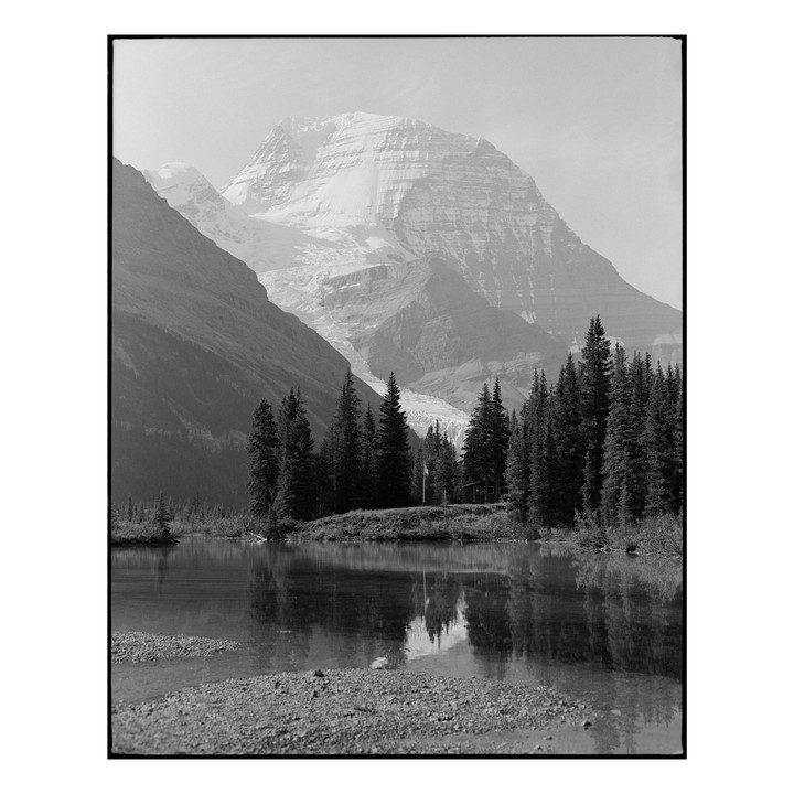 A snow-capped mountain rises behind a forest and a small cabin, with a lake in the foreground.
