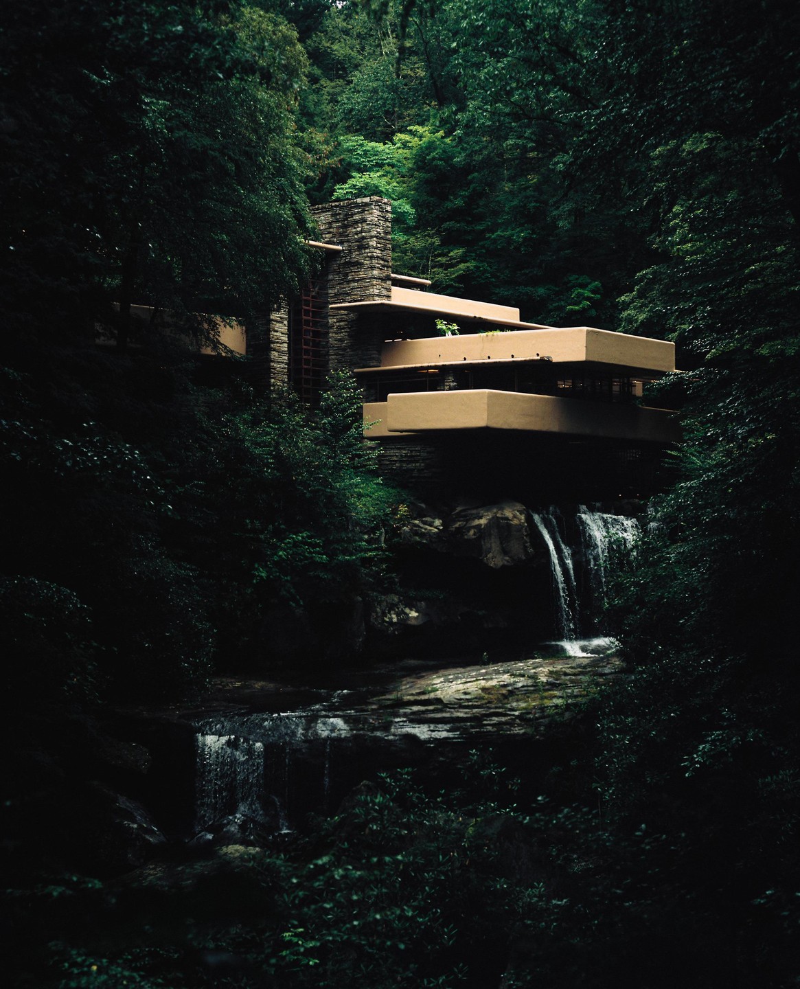 A modern house with multiple balconies is built into a lush, green forest above a waterfall.