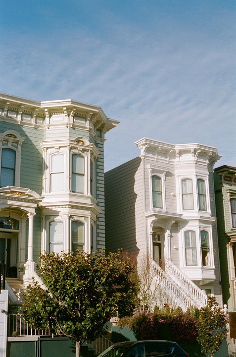 Three Victorian houses with bay windows and ornate trim stand under a clear sky, with a dark car visible at the bottom.