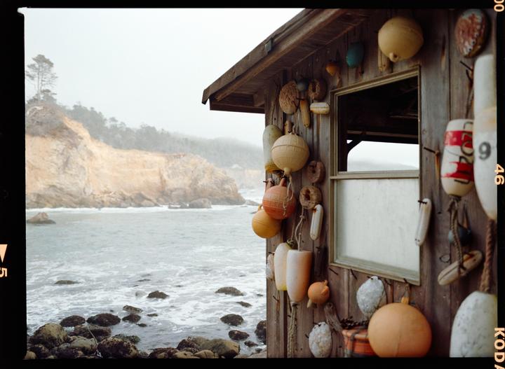 A wooden shelter adorned with buoys stands beside a rocky coastline with a cliff in the background.