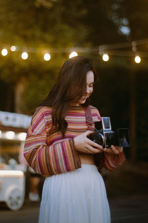 A woman in a striped sweater and white skirt holds a camera under string lights.