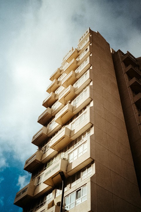 A tall concrete building with many balconies is photographed from a low angle against a bright sky.