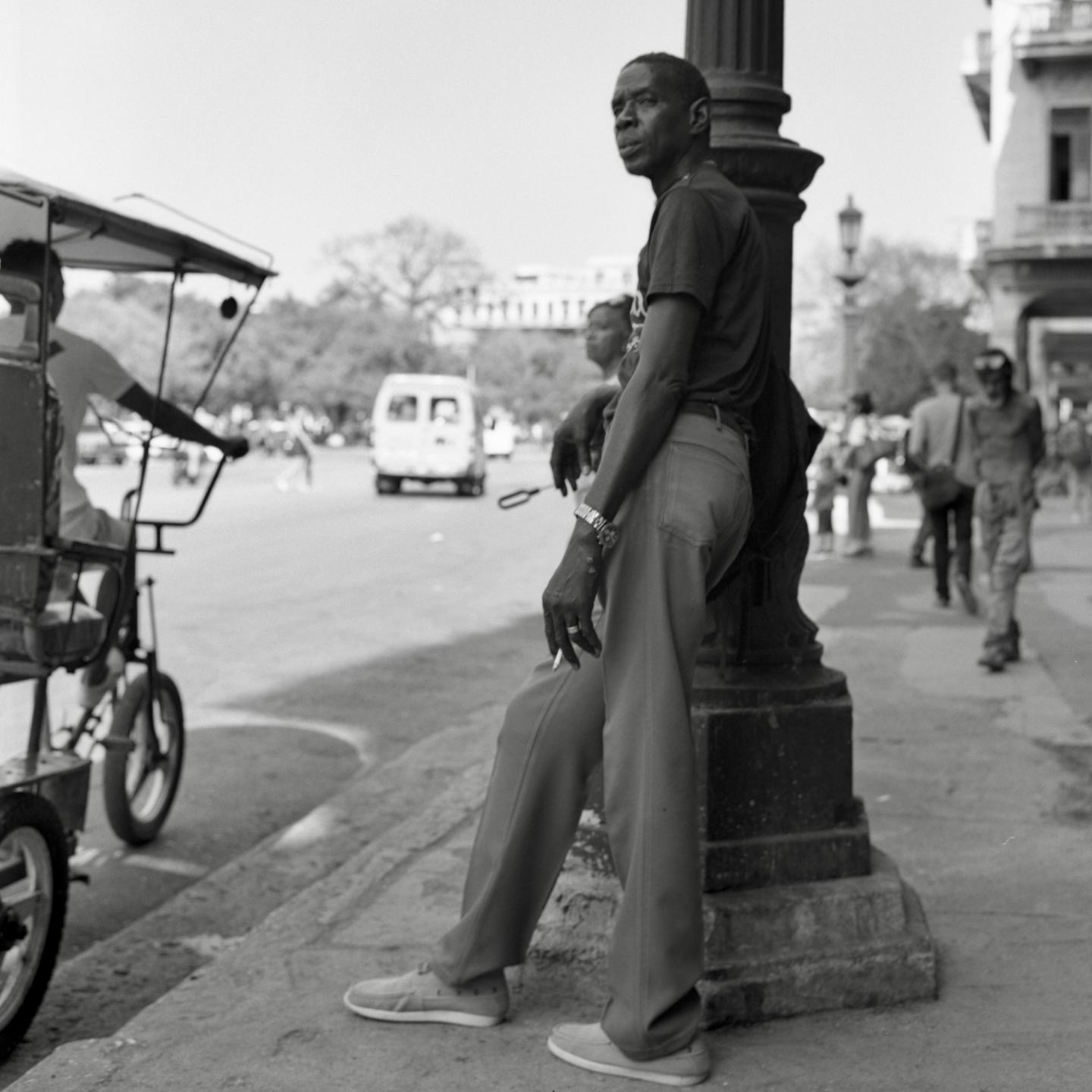 A man leans against a lamppost on a street in Havana, Cuba, with a cigarette between his fingers.