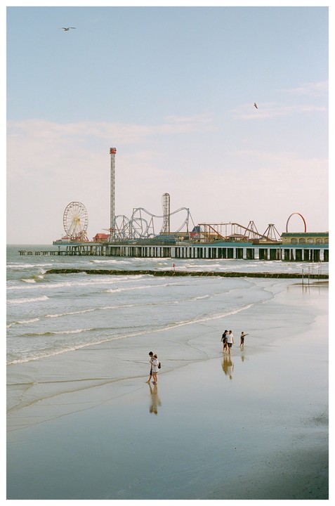 People walk on a wet beach with an amusement pier in the background.