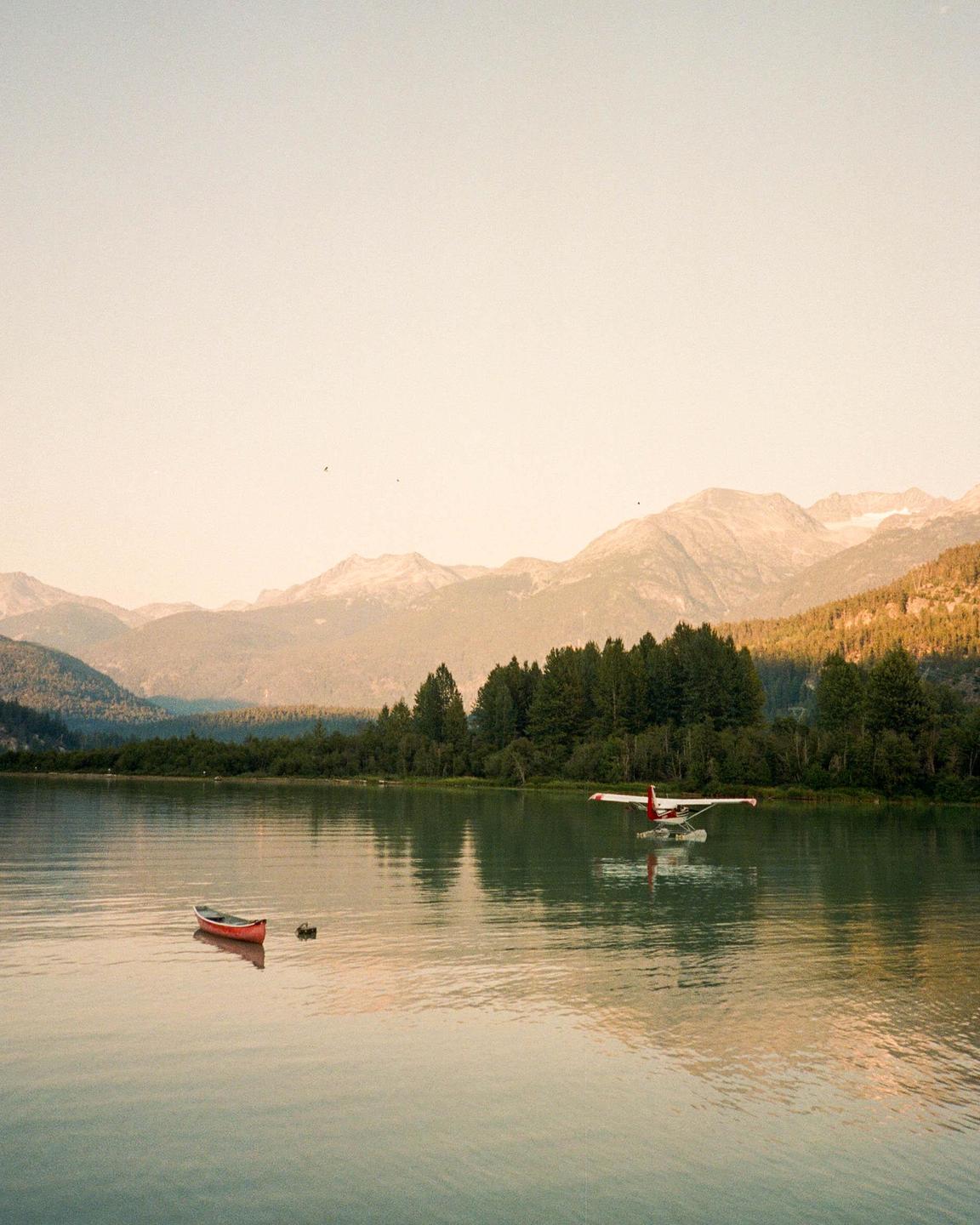 A red canoe and a seaplane are on a lake surrounded by mountains and forest.