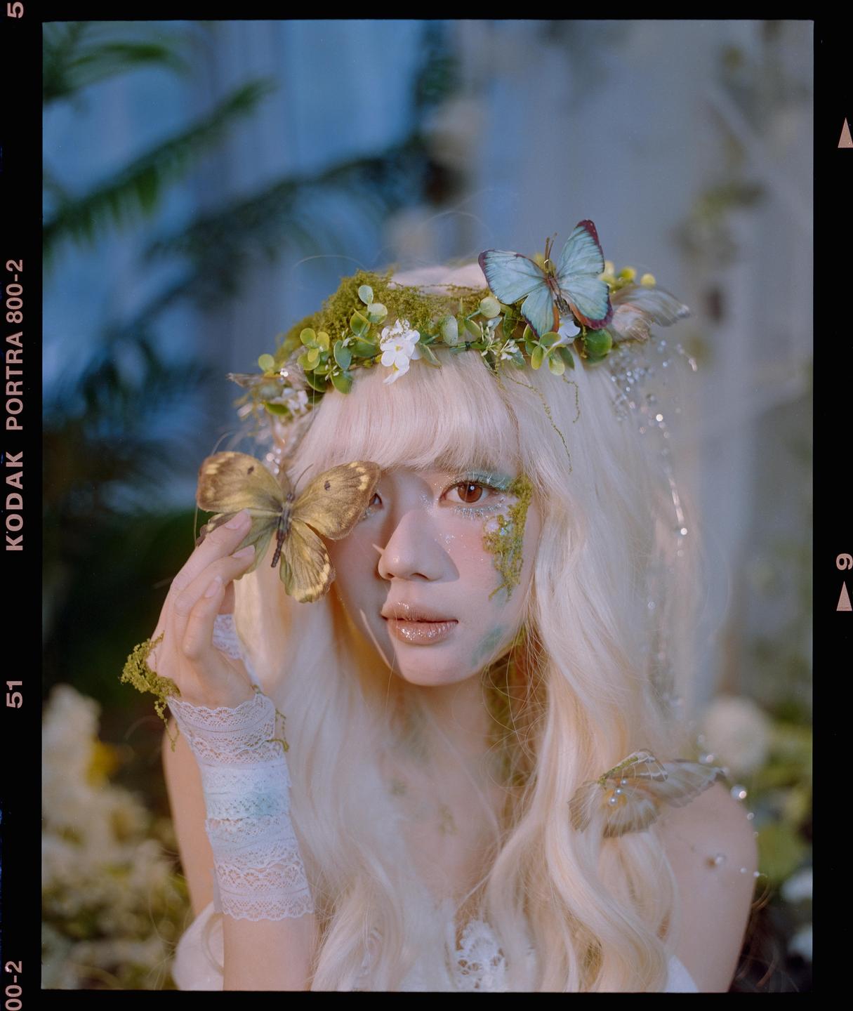 A woman with long blonde hair and a floral crown holds a butterfly near her face, which is adorned with glitter and moss.