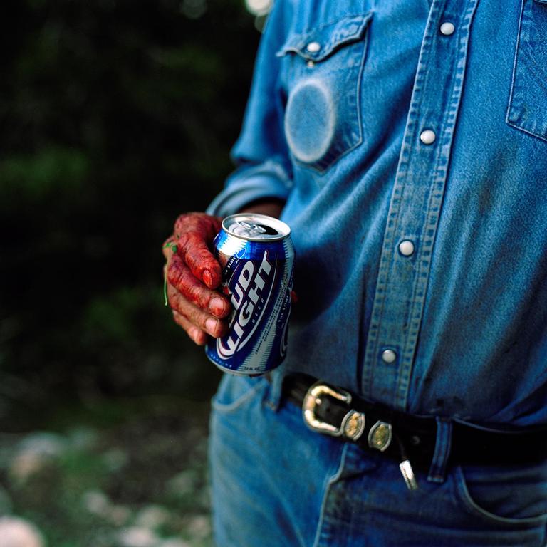 A person in a denim shirt and jeans holds a can of Bud Light with a blood-stained hand.