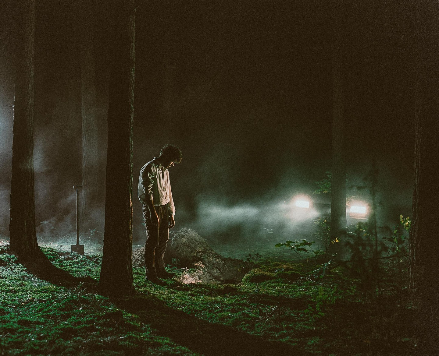 A man stands in a dark forest at night with a shovel nearby and car headlights in the distance.