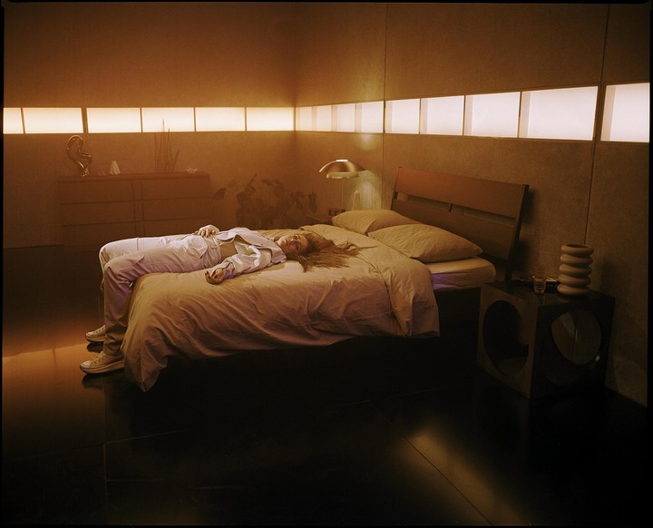 A woman with long hair lies on a bed in a dimly lit room with glowing rectangular lights on the wall.
