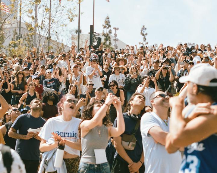 A large crowd of people are gathered outdoors, many with their hands raised, looking up at something out of frame.