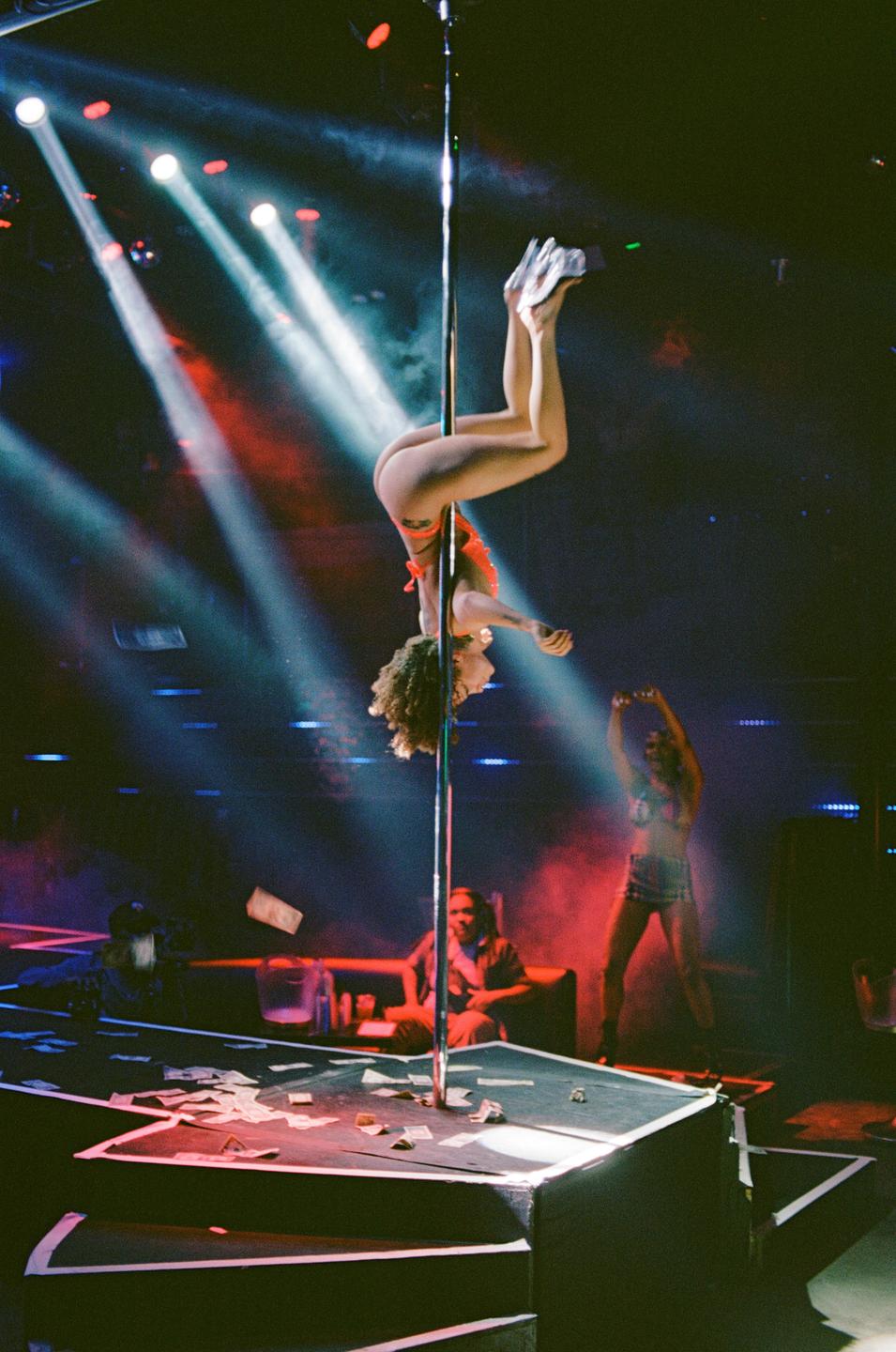 A woman hangs upside down from a pole on a stage with money scattered on it, while other people are visible in the background.