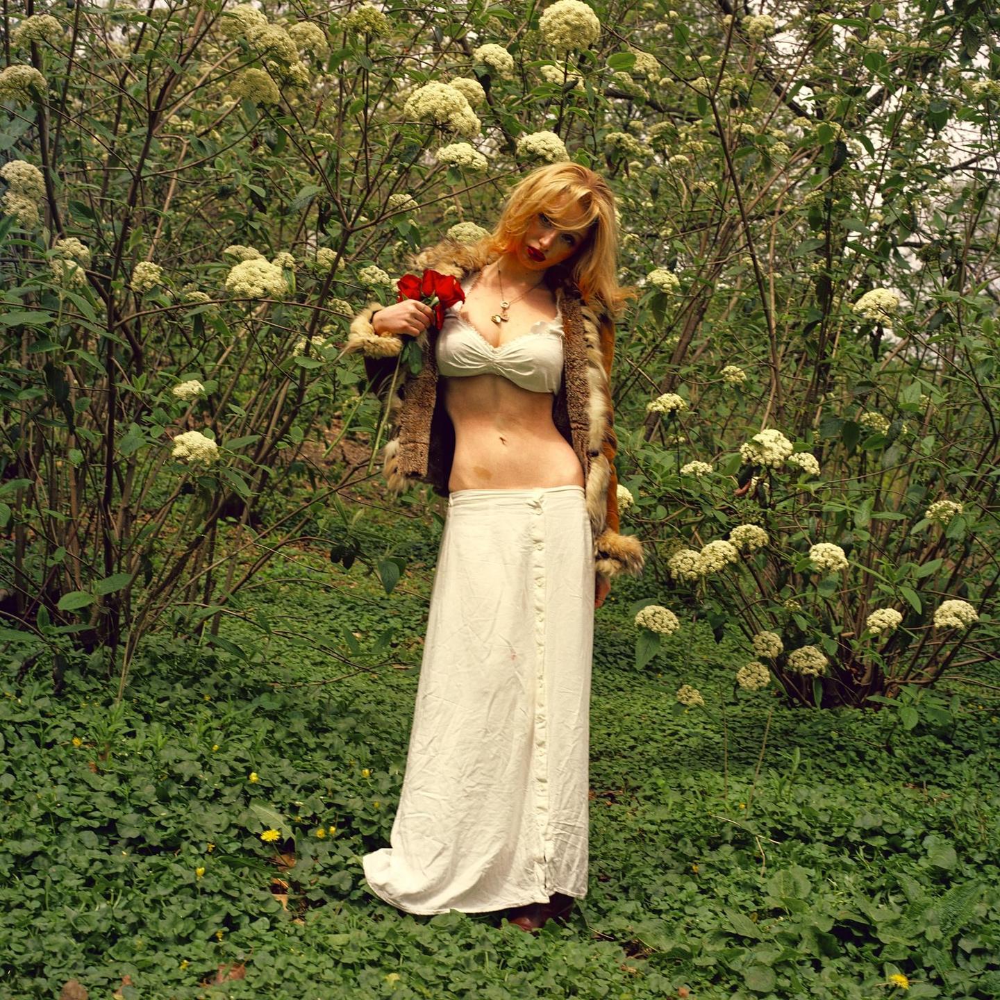 A woman wearing a fur-trimmed jacket and a long white skirt stands in a garden holding red roses.