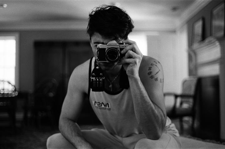 A man in a tank top holds a camera to his face, capturing his reflection in a self-portrait.