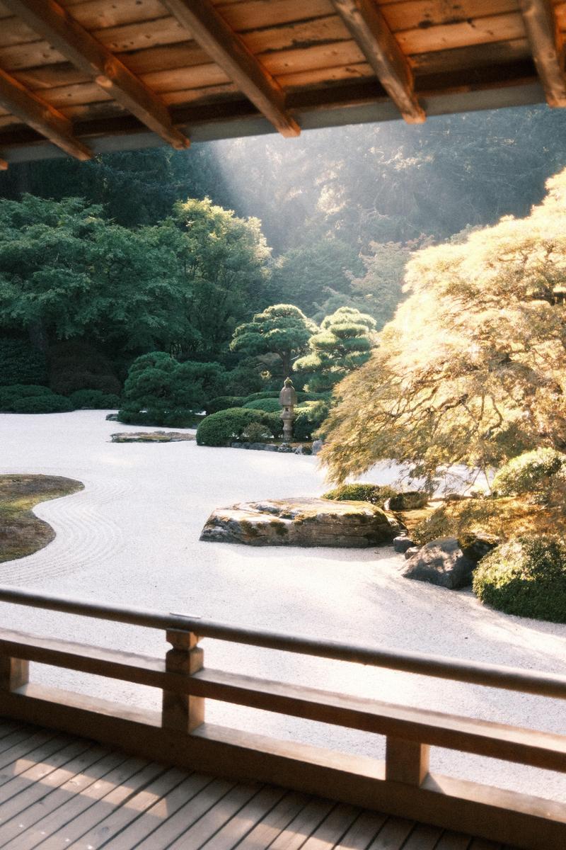 A zen garden with raked sand, rocks, and trees is viewed from under a wooden roof and balcony.