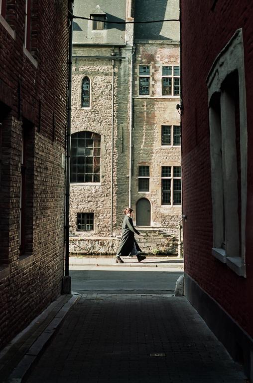 A woman in a long coat walks along a narrow street between brick buildings.