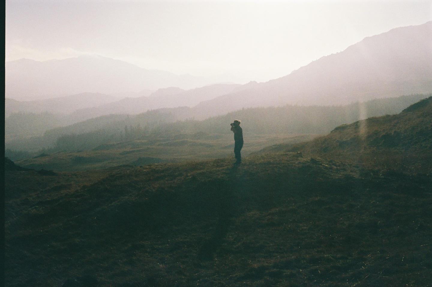 A person stands on a grassy hill, looking out at hazy mountains in the distance.