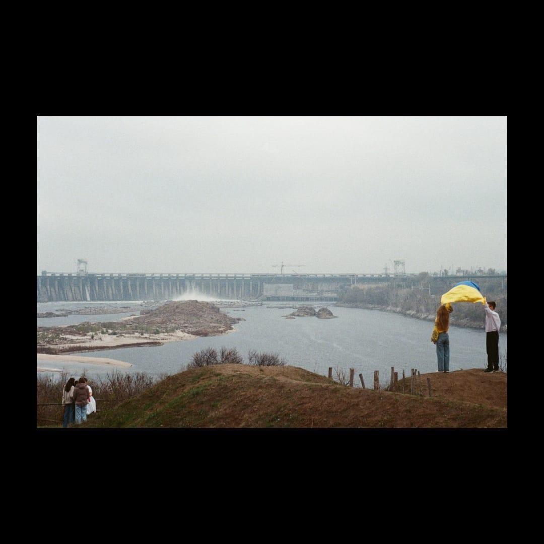 A couple holds a Ukrainian flag on a hill overlooking a river and a large dam under a hazy sky.