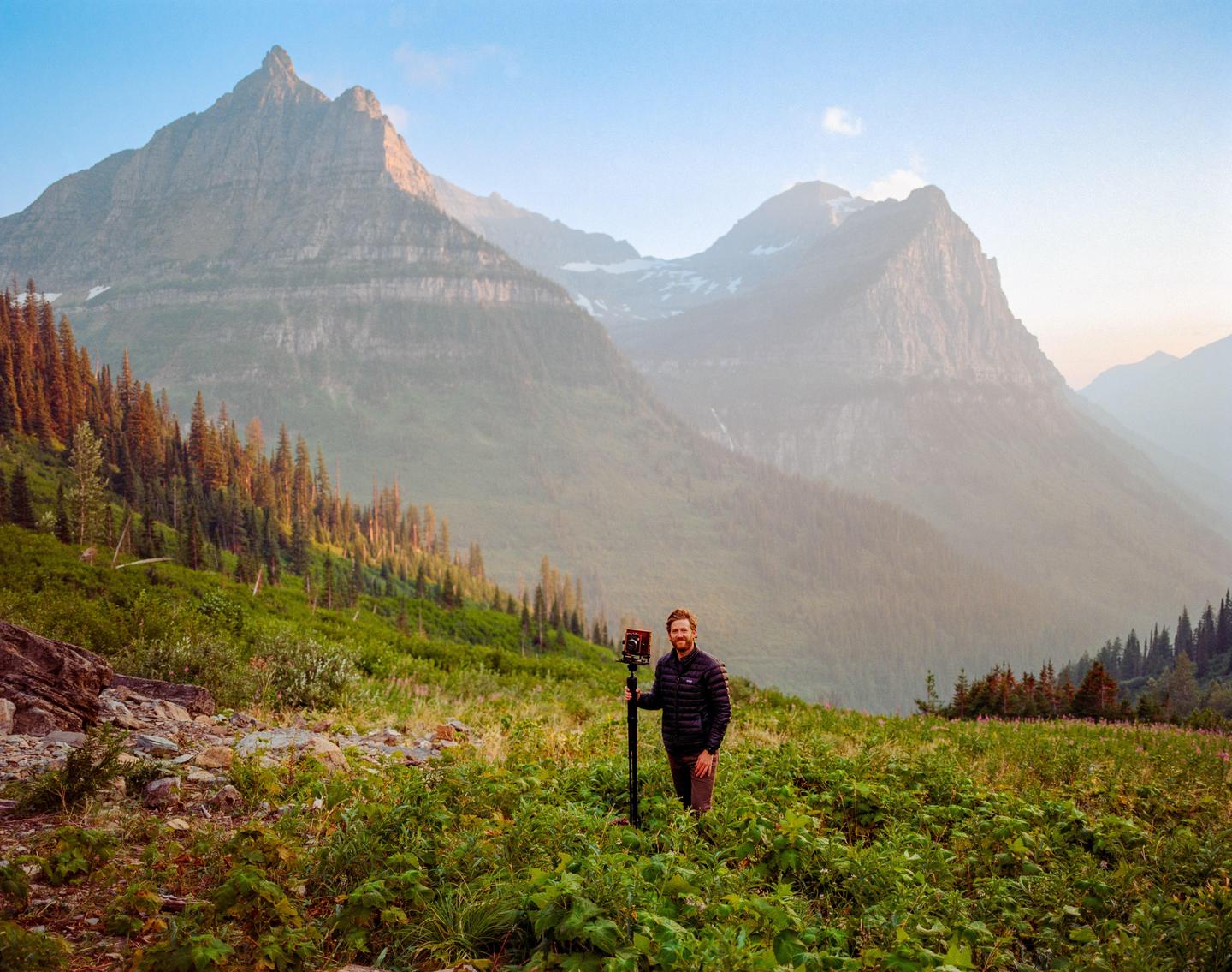 A man stands with a camera on a tripod in a field of green plants with mountains and trees in the background.