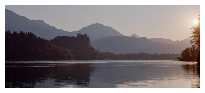 A panoramic landscape shows a lake with a castle on a hill, surrounded by mountains under a hazy sky at dawn.