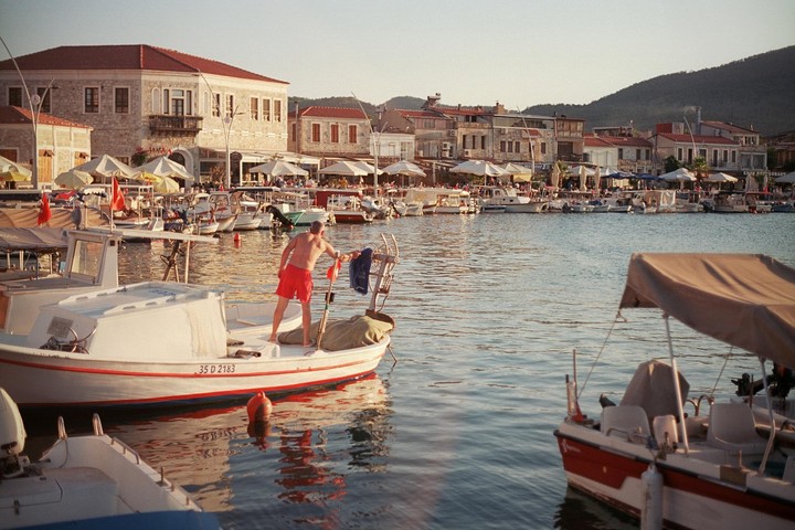 A man in red shorts stands on a boat in a harbor with buildings and other boats in the background.