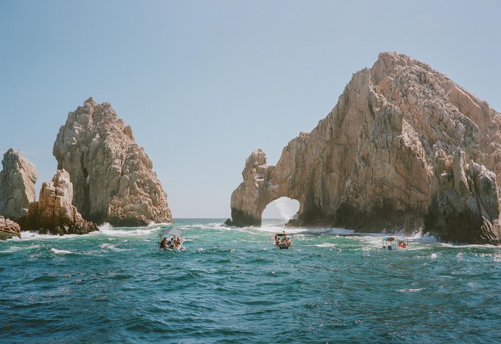 Three small boats with people are on the water near large rock formations, one of which has a natural arch.