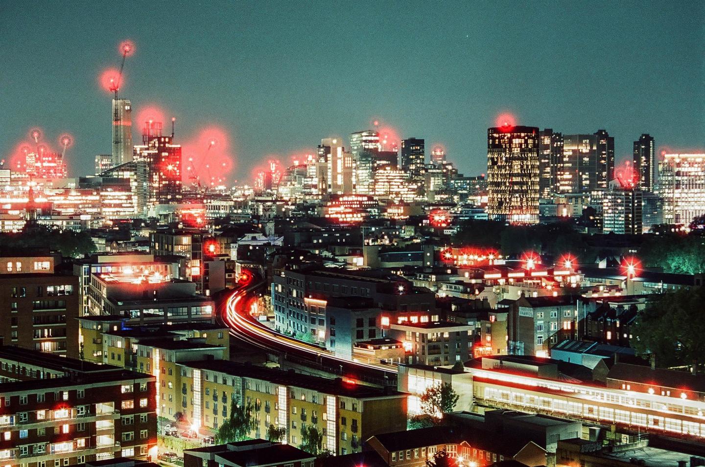 A cityscape at night shows buildings with illuminated windows and red lights on top of skyscrapers, with light trails from moving vehicles in the foreground.