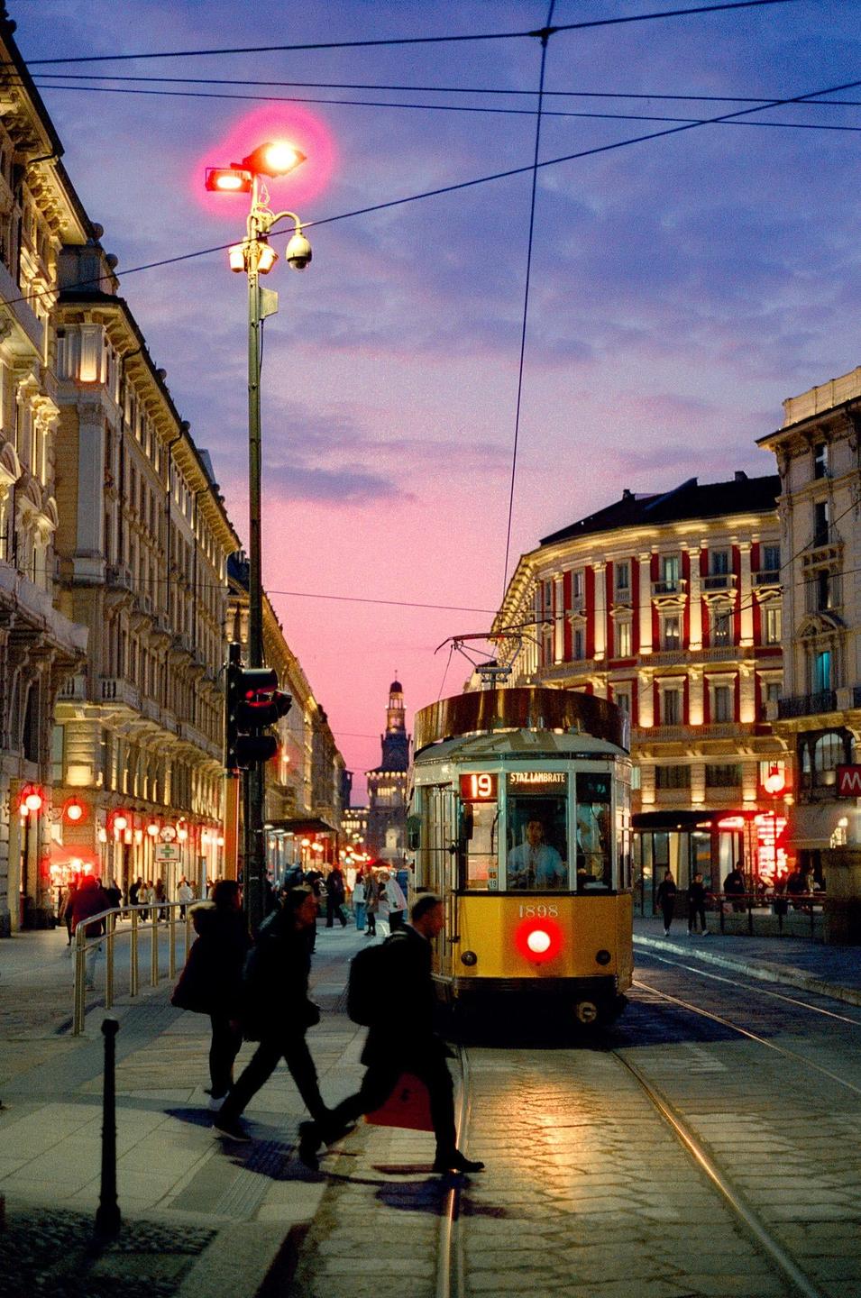 A yellow tram is stopped on a city street with people walking by and buildings lit up under a pink and purple sky.